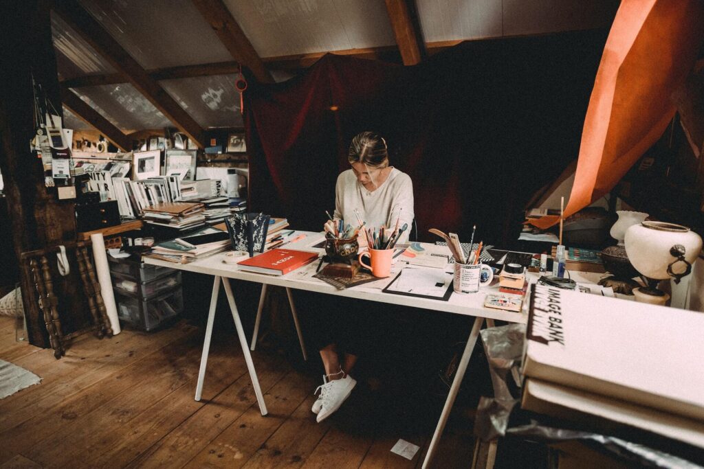 Artist in a makeshift studio, sitting on a table drawing, surrounded by art books, art tools.