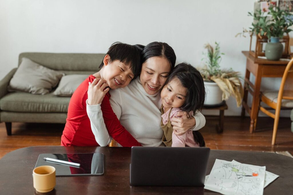 Woman hugging children while sitting down in front of desk with an open laptop and a tablet