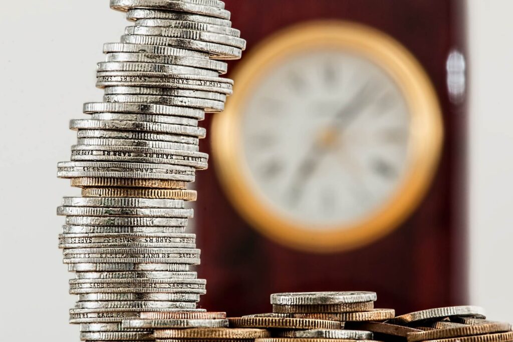 Stack of coins on the forefront with a blurred clock in the background
