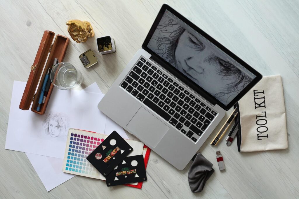 Laptop with a drawing on the screen, surrounded by artist's tools, paper with drawing, color swatches