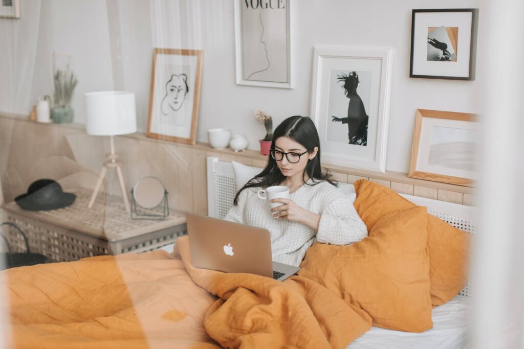 Woman wearing glasses in bed, using a laptop, holding a mug