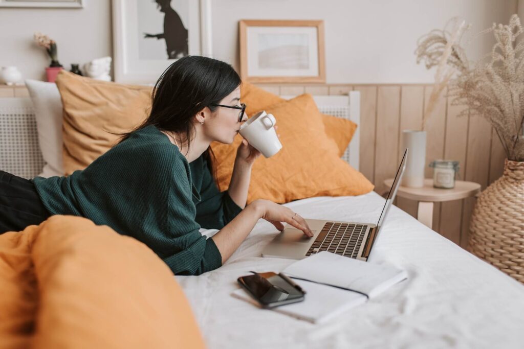 Woman lying on her stomach, using a laptop on her bed, while drinking coffee