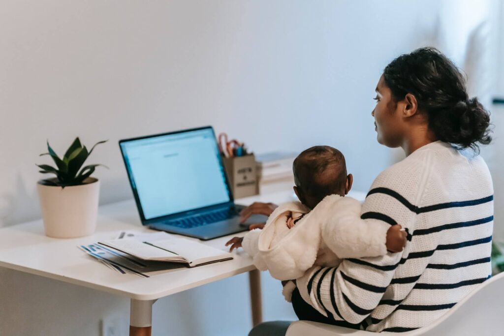 Mother holding a baby while sitting in front of a desk, working on a laptop