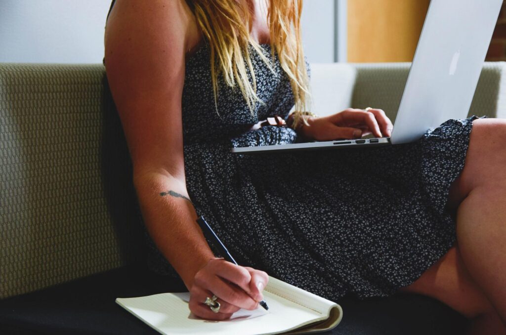 Woman using a laptop while writing on a notebook with a pen.