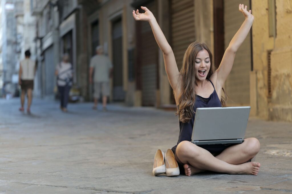 woman sitting on street with arms raised and looking at laptop