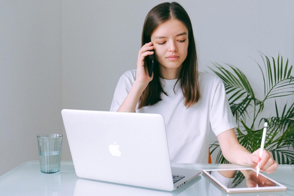 Woman on the phone sitting in front of a laptop while using a pen and tablet