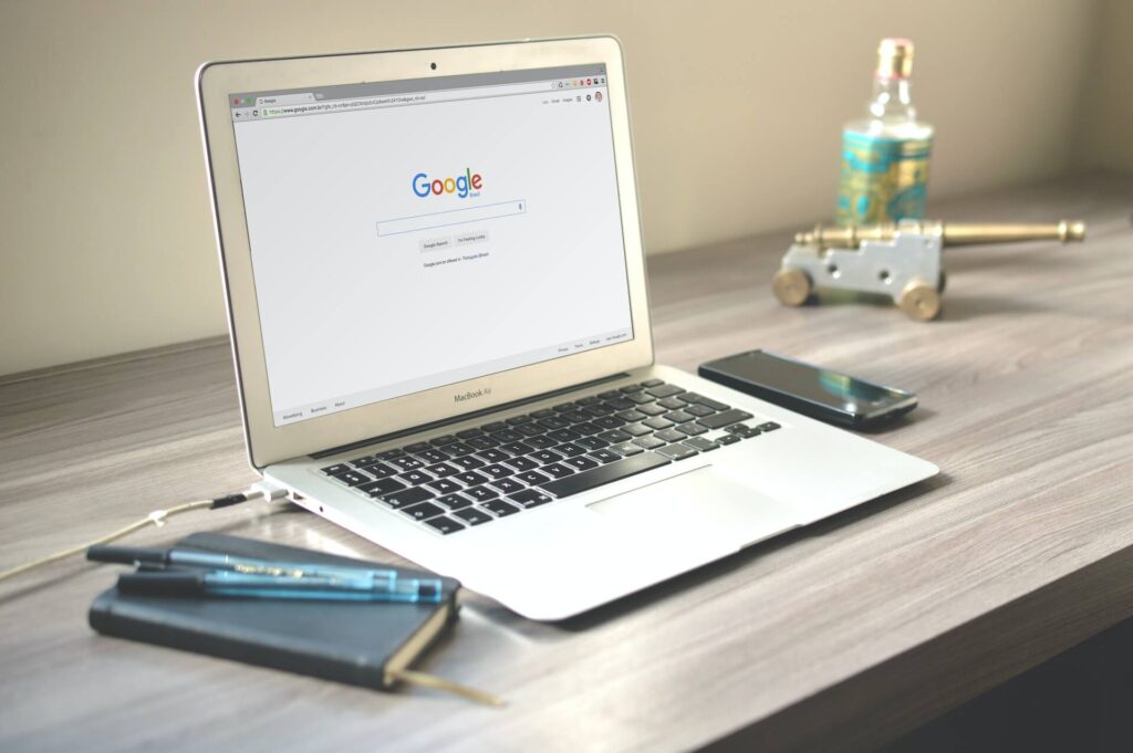 Desk with a mobile phone, notebook, pens, and an open laptop showing the Google home page. 