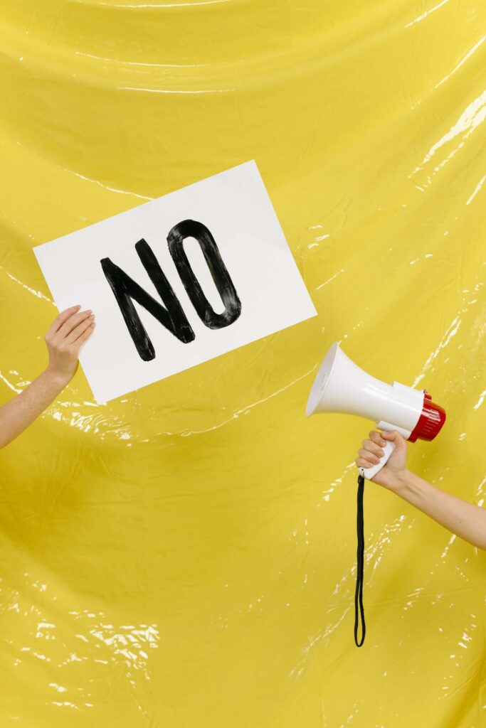 Yellow background, hand holding a placard that says No, and another hand holding a megaphone