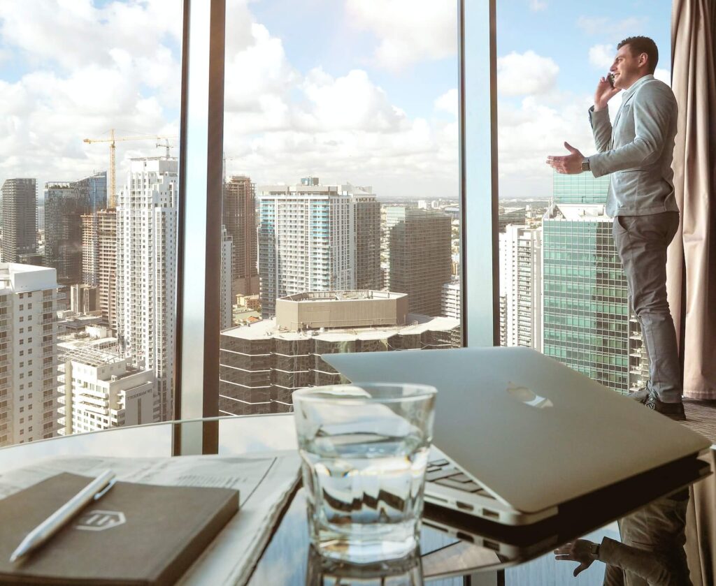 Man talking on the phone, standing in front of a window showing surrounding buildings. On the foreground, a table with a glass, laptop, pen, notebook, and newspaper.