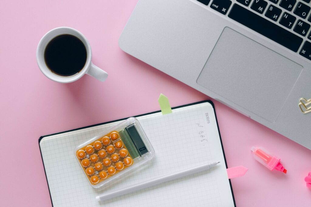 partial laptop, calculator, coffee, notebook and pen on a pink table