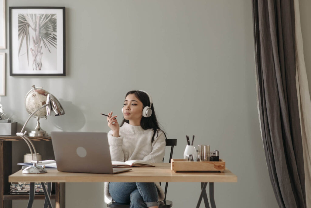 Woman wearing headphones holding a pencil, sitting on a chair in front of a desk with a laptop