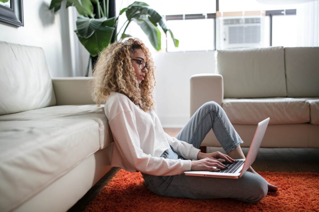 Woman sitting on a carpet on the floor, back to the sofa, using a laptop