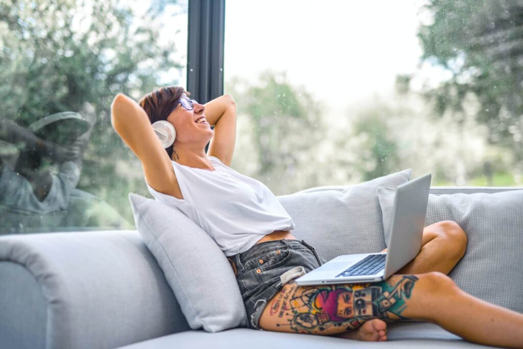 Woman smiling, sitting on couch with a laptop on her lap, both hands behind her head