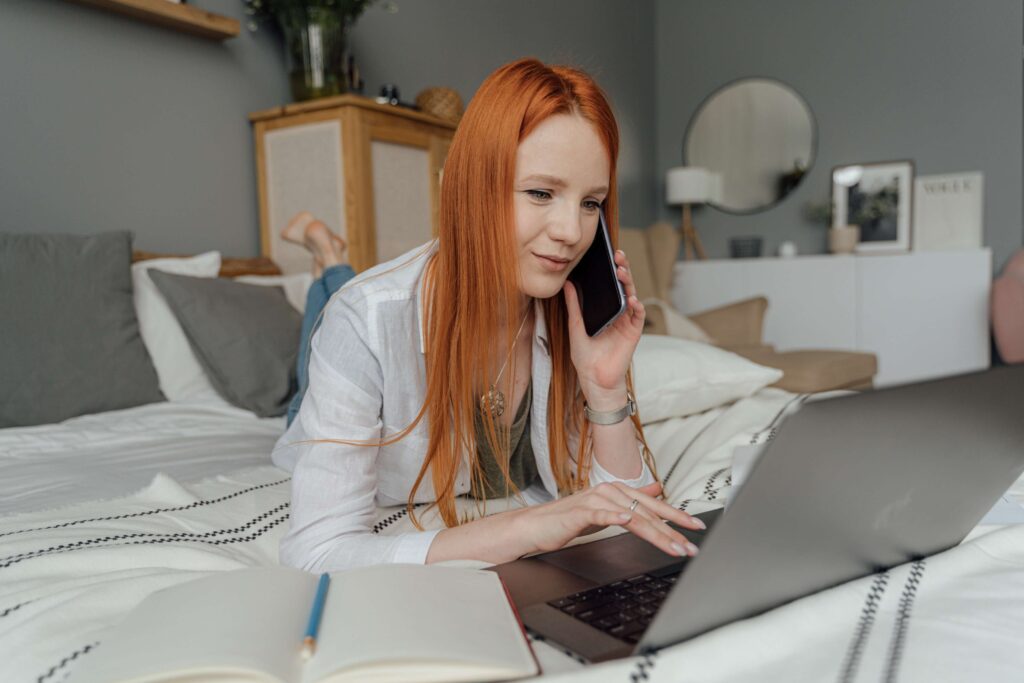 Woman using laptop on bed while on the phone