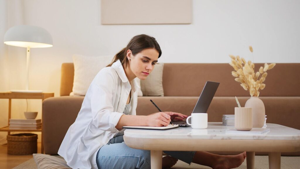 Woman sitting on the floor, working on laptop and writing on a notebook with a pencil, both placed on top of a coffee table.