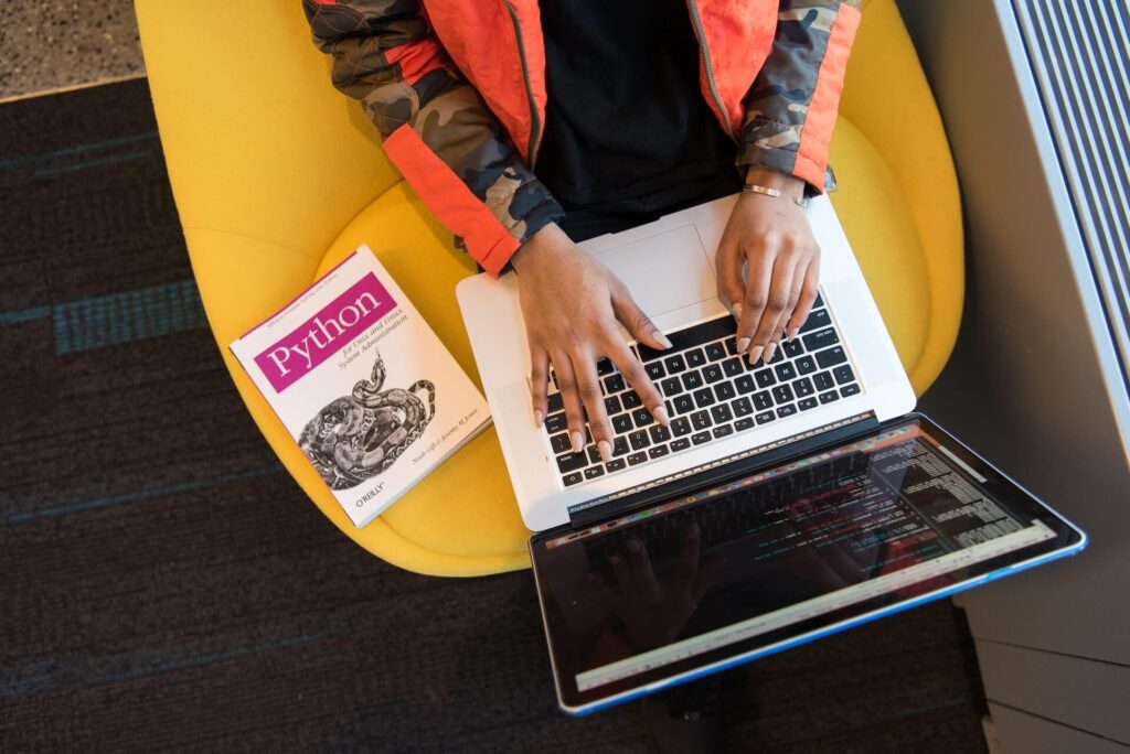 Top shot of woman using a laptop, a Python textbook beside her