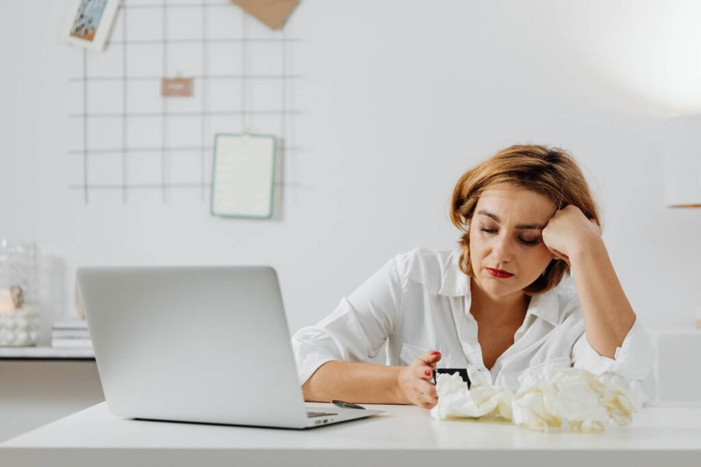 Woman looking at phone resting arm on table with a laptop and crumpled up tissue