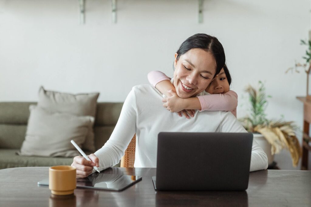 Woman working using a laptop and tablet while her child hugs her from behind