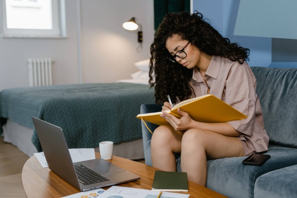 Woman sitting on couch writing on notebook, laptop on coffee table in front of her