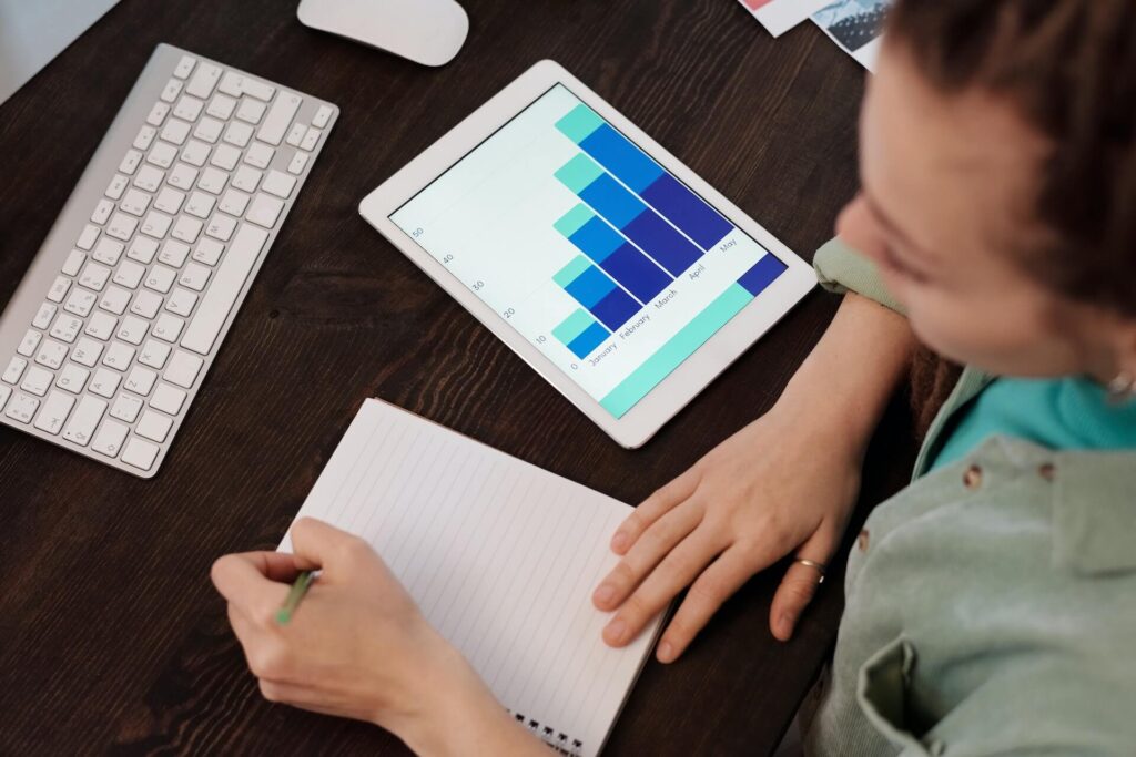 woman writing on notebook with chart on tablet screen