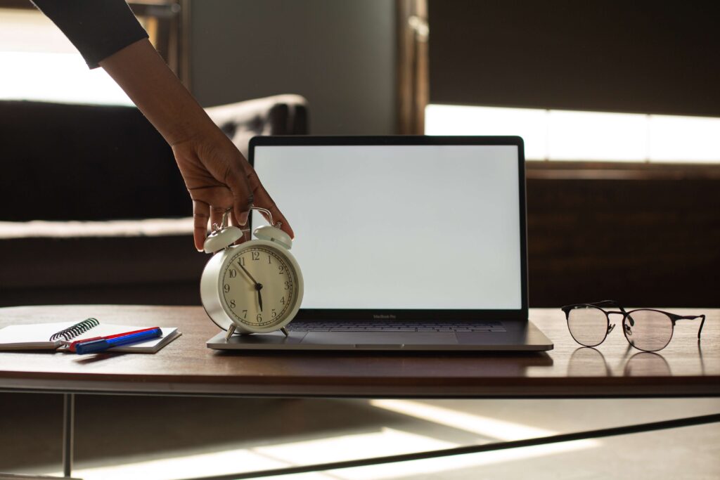 Table with a laptop, eyeglasses, notebook, pens, and an alarm clock