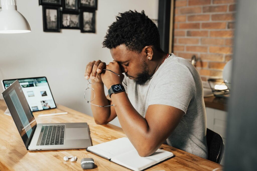 Man looking tired holding eye glasses, working on a desk with a laptop, iPad, and notebook on top of it. 